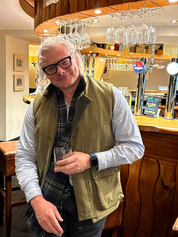 Peter standing against a bar, sipping scotch in a Gilet taken whilst on Holiday in April 2025